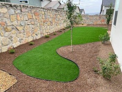 Artificial turf with organic S-curve, young tree, and bark mulch beside a stone wall