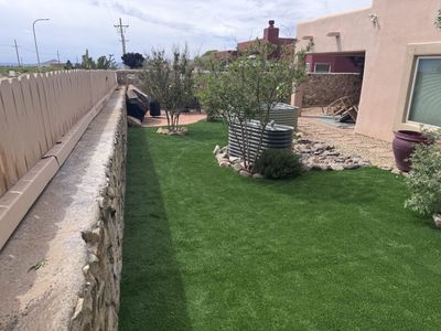 Side yard artificial turf with stone retaining wall and galvanized stock-tank planters beside an adobe home