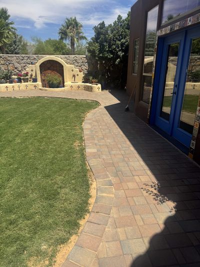 Brick paver walkway curving past green lawn to a Talavera-tiled stucco arch wall with blue french doors