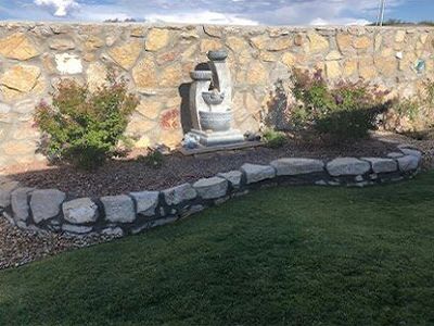 Tiered fountain with stone border, flowering plants, and turf lawn against a stone wall