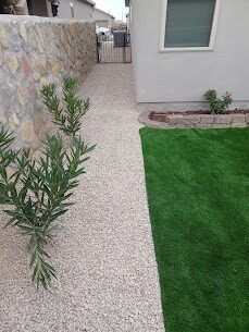 Side-yard artificial turf with olive tree and gravel border along a stone wall